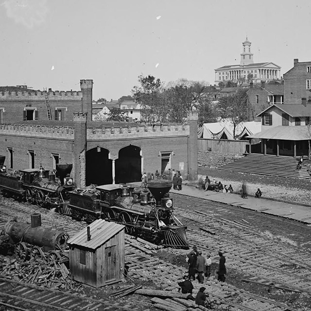 Steam locomotives sit on railroad tracks in front of brick buildings.