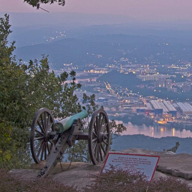 Two cannons overlook the city of Chattanooga at dusk with trees framing the picture.