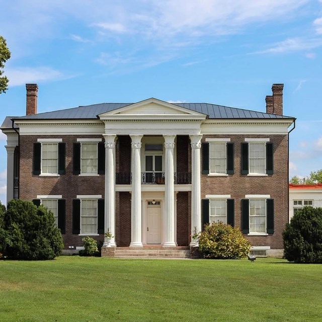 A two story brick house with tall glass windows with 3 chimneys and four columns in front of it.