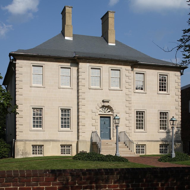 A three-storied white house with two chimneys is surrounded by a grassy lawn and stone fence