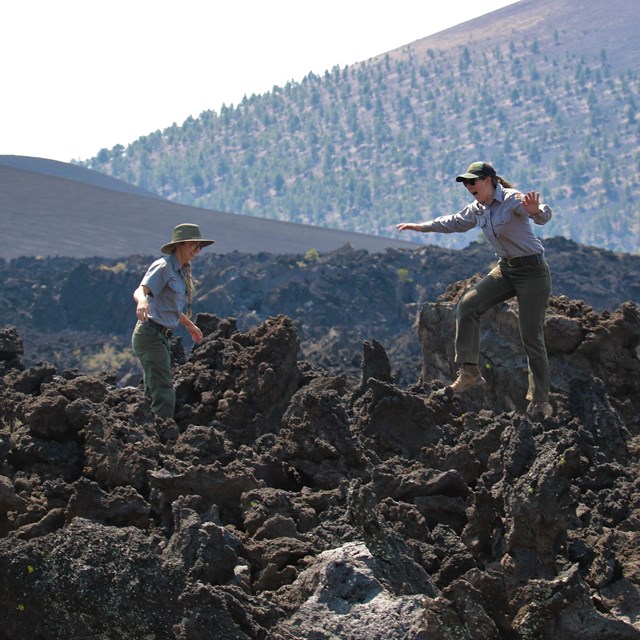 two park rangers standing atop a jagged lava flow with a cinder cone in the background