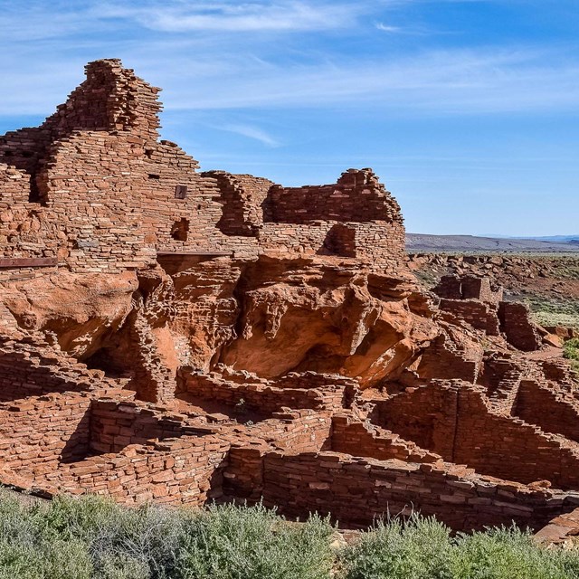 Red rock walls of an ancestral village stand in green grass under a blue sky