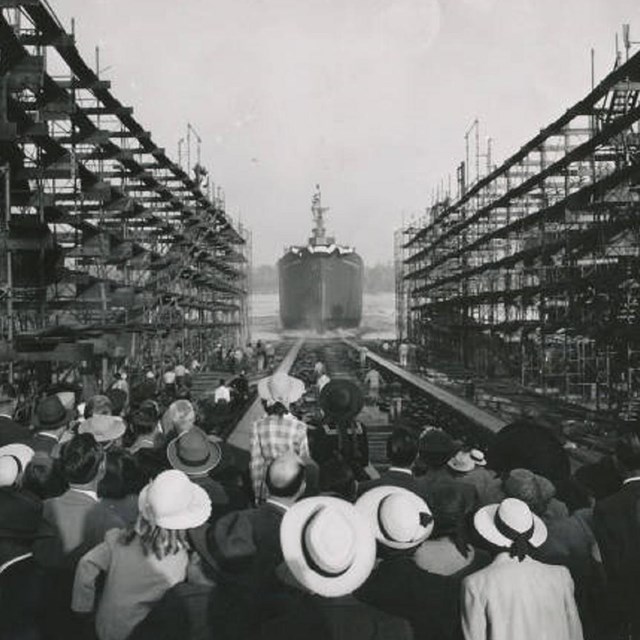 Dozens of people watch as a ship launches into the ocean from a marina.