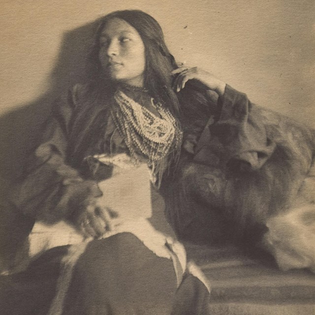 A seated woman with long hair wearing beads looks to her right. She wears a long flowing robe.