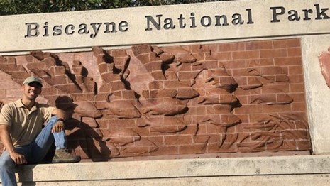 A brown male posing in front of the Biscayne National Park sign entrance