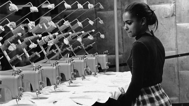 A young African American woman watches at least seven sewing machines working simultaneously.t