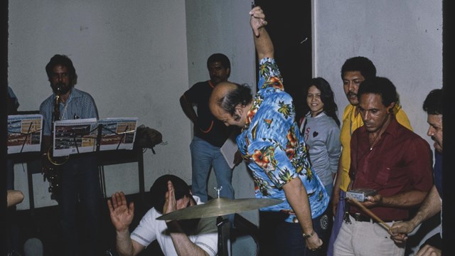 Salsa band in rehearsal with eight people in the frame. Man in the middle is wearing a blue shirt