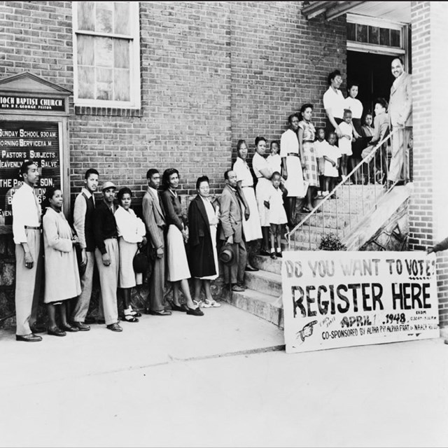African Americans stand in a queue on church stairs behind a sign saying register here