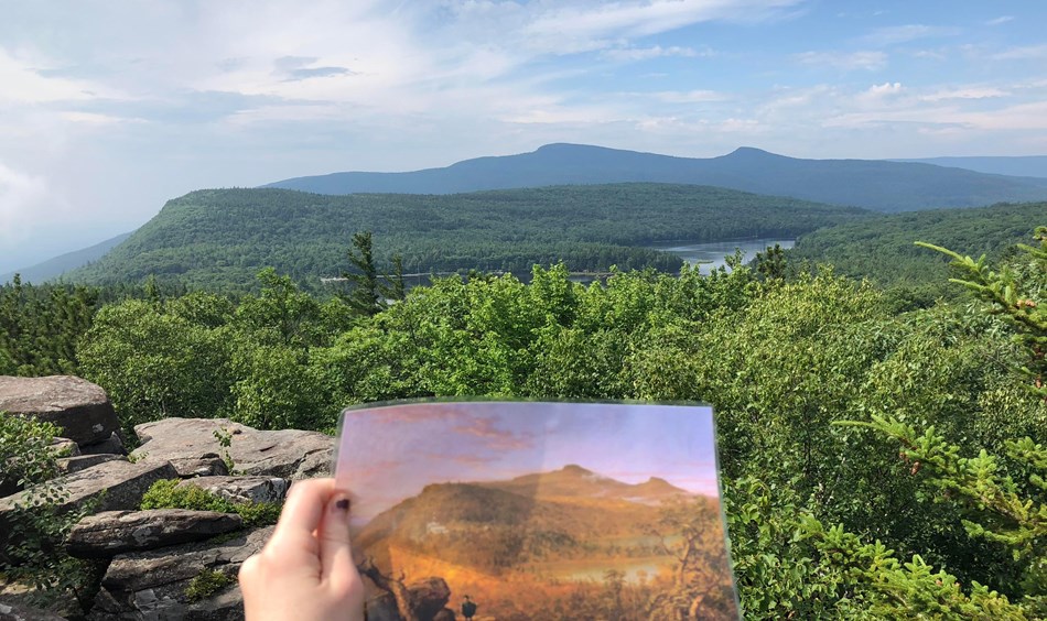 A view of a distant mountain in shades of blue & green. A hand holds a portrait image of the same.