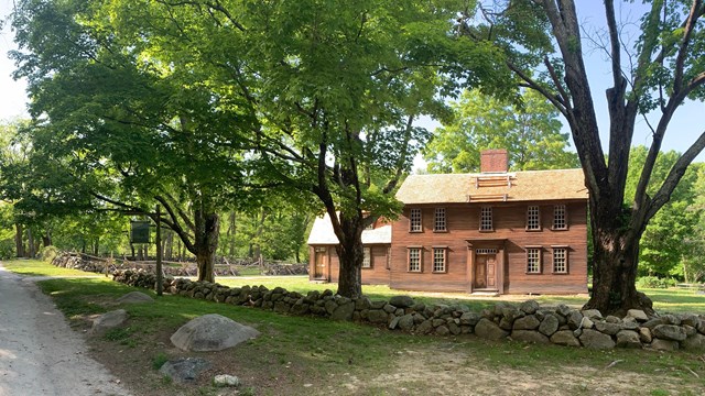 A wooden colonial house sits along a dirt road surrounded by trees and stone walls.