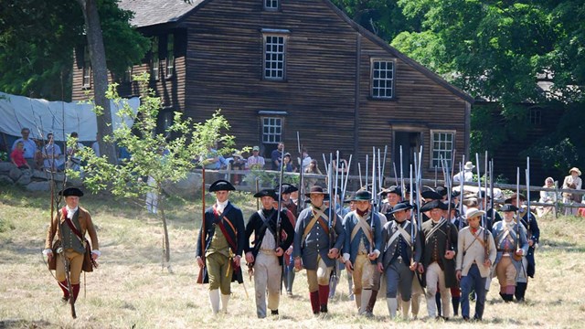 A wooden colonial house in the background. A formation of colonial militia soldiers marches in front
