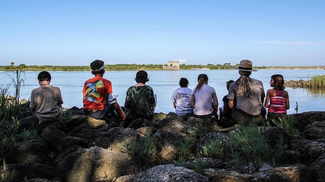 7 kids and 1 park ranger sitting on rocks.  Fort in background.