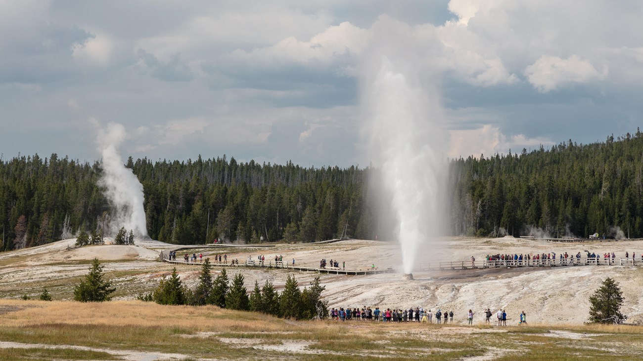 People stand watching erupting geysers from the safety of a boardwalk.