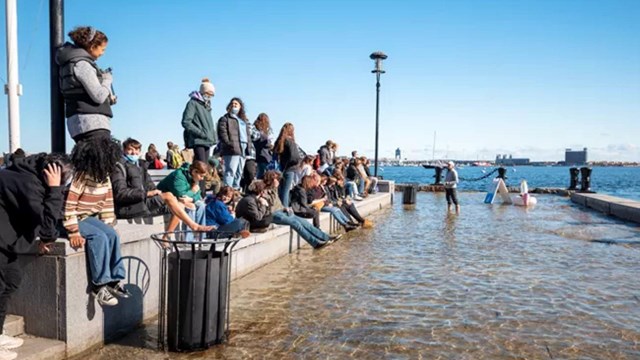 A crowd at the edge of a harbor with someone speaking to them standing in high water