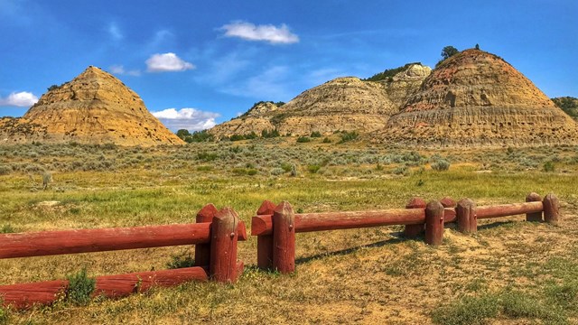 Brown fence post in front of green grass, buttes, and blue sky overhead