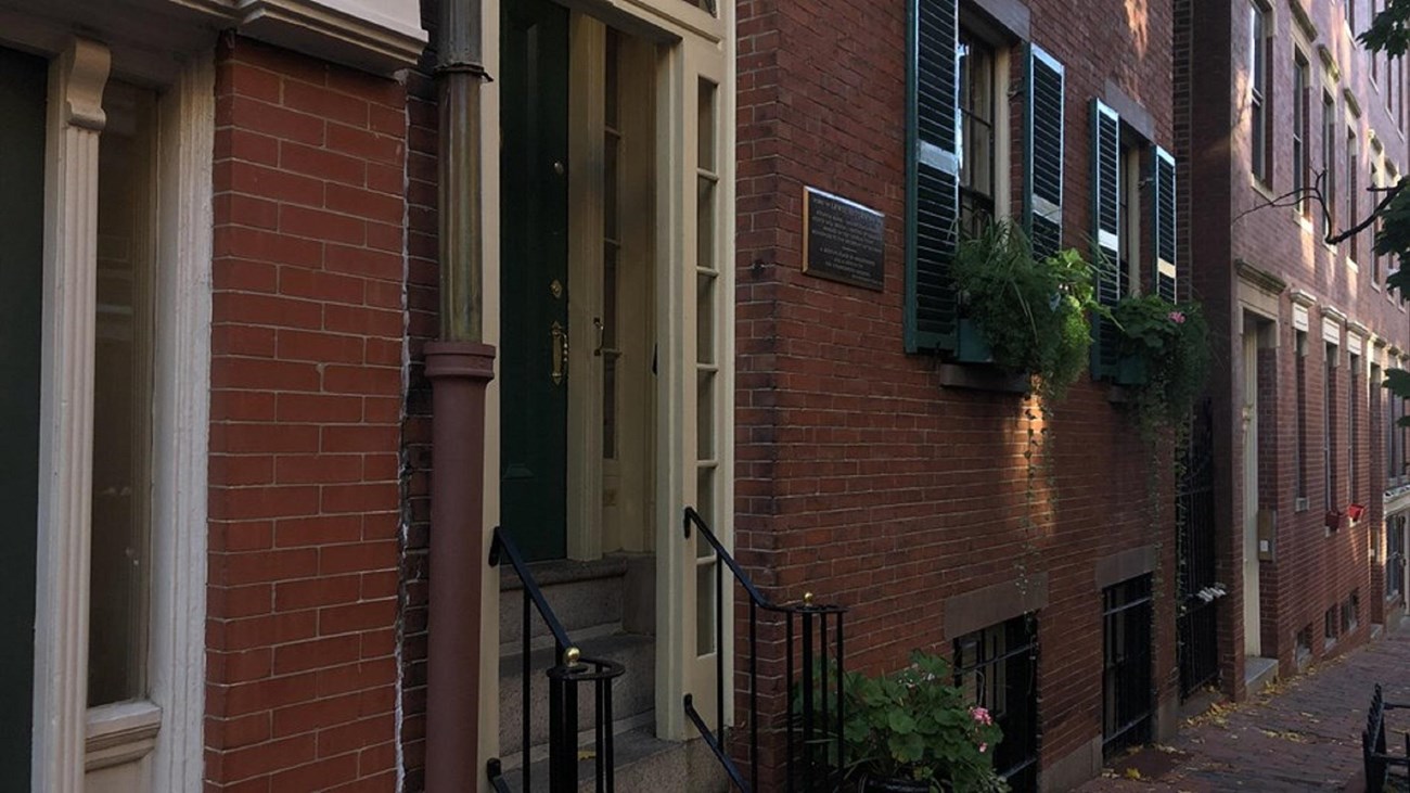 Photograph of a brick building from an angle to include the front door, windows, and sidewalk.