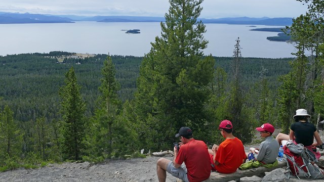 Hikers rest and look out at Yellowstone Lake from atop a mountaintop.