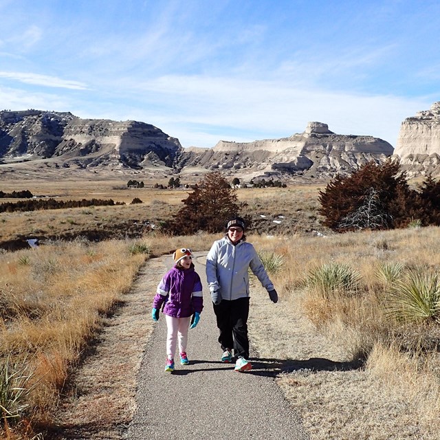 A pair of hikers walks a trail with dramatic rock formations in the background.