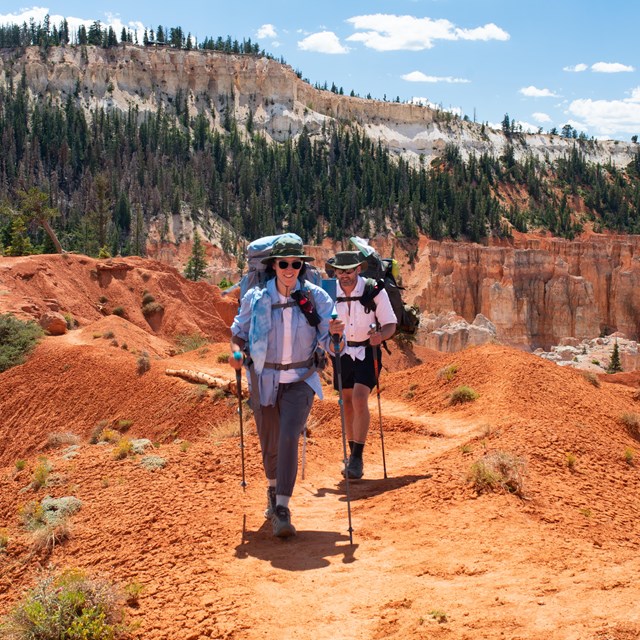 Two hikers carry their gear as they hike along a dirt trail.