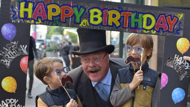A man dressed as Theodore Roosevelt is posing with a frame with two boys, also dressed as TR