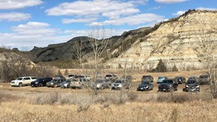 Twenty vehicles parked in a gravel lot at a trail head surrounded by trees and layered buttes.