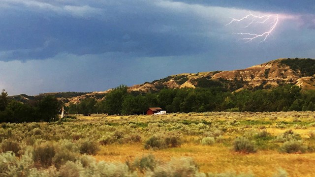 Lightning crackles from behind a dark blue cloud atop an open prairie with a butte behind.