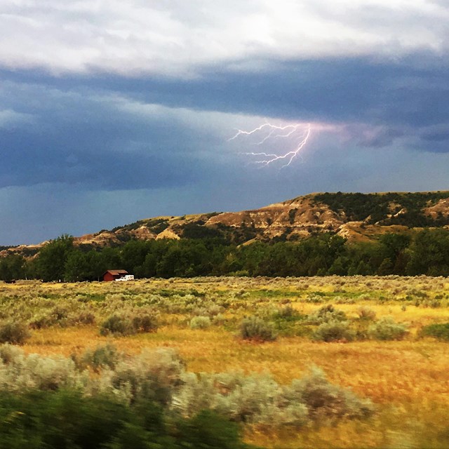 Lightning crackles from behind a dark blue cloud atop an open prairie with a butte behind.