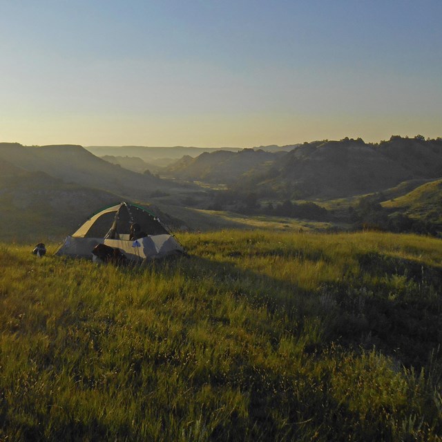 Sun floods a scene with a small tent in a green field with badlands buttes beyond