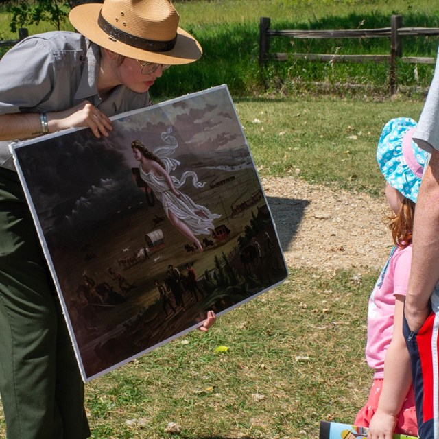 A ranger shows a large photo of a painting to a group of visitors.