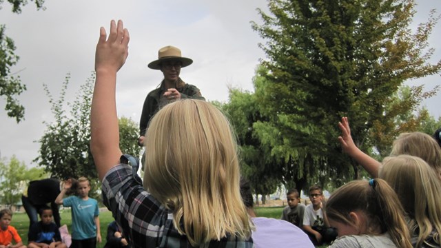 Multiple children with hands raised to ask a question sit around a ranger
