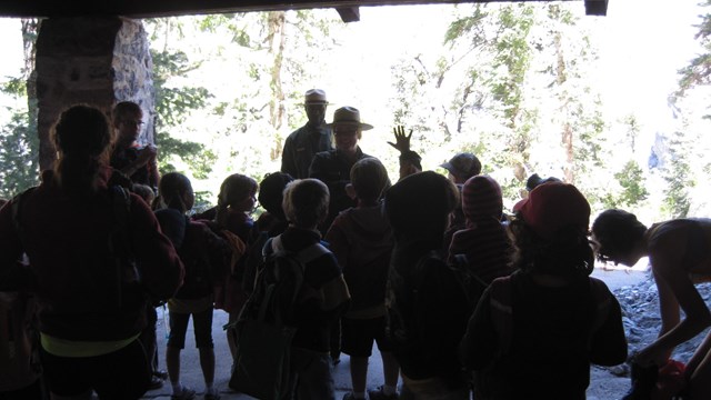 A ranger backlit by the natural light with multiple school children gathered in front.