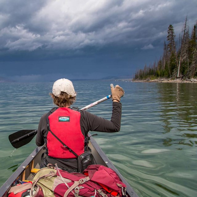 a boy in a red lifejacket paddles a canoe on a lake