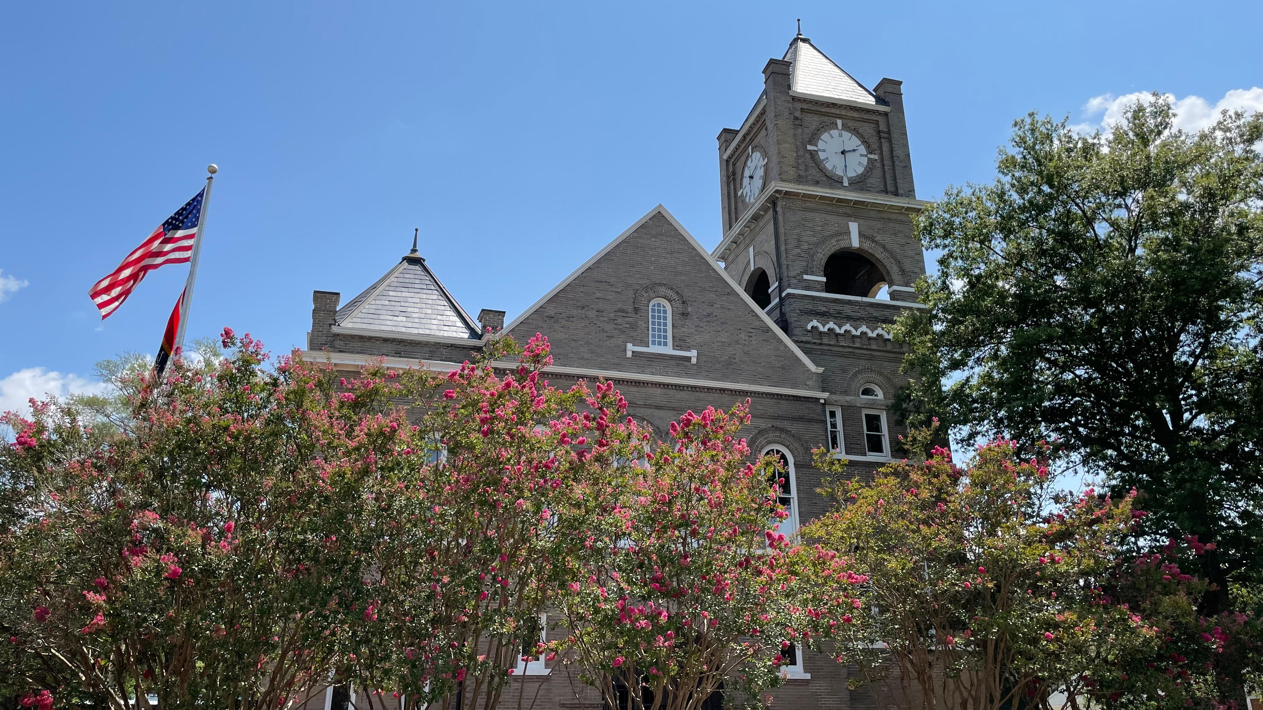 A two-story brick courthouse with clock tower and trees in front.
