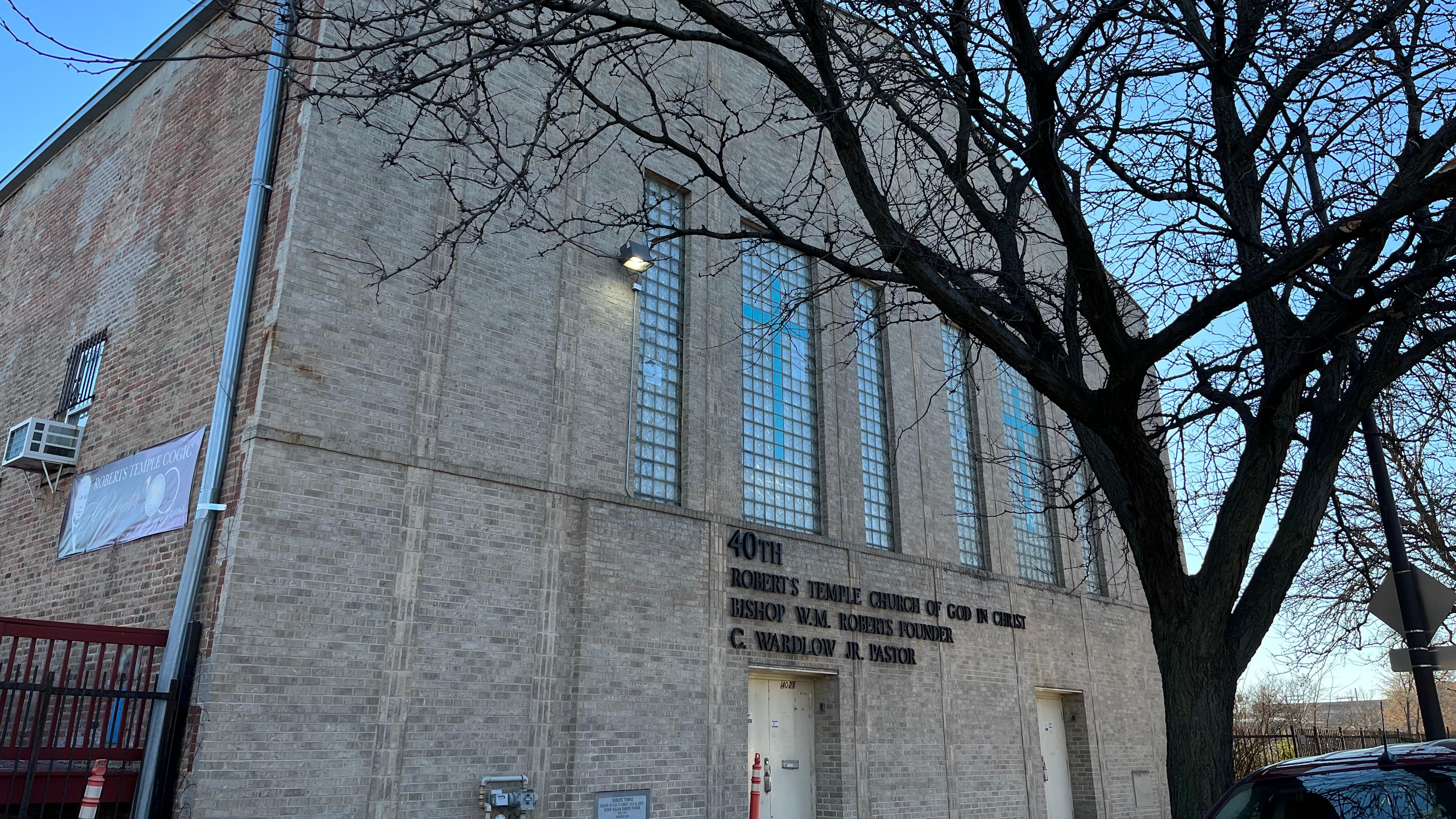 Front corner of a multi-story tan brick church with a tree in front at dusk. 