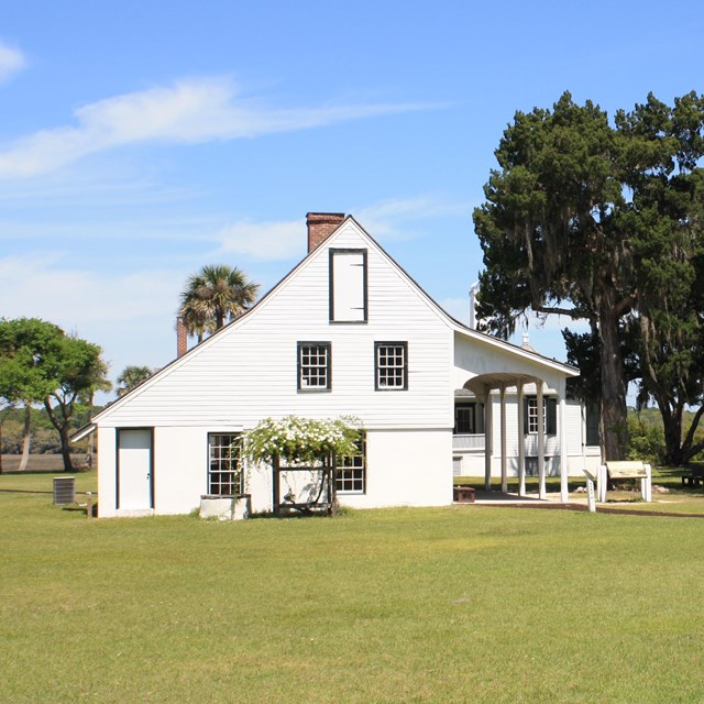 the white kitchen house with lush green grass and trees surrounding 