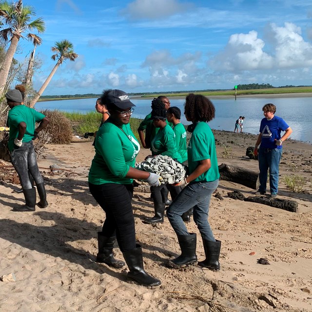 young people in green shirts carry oyster forms on beach  