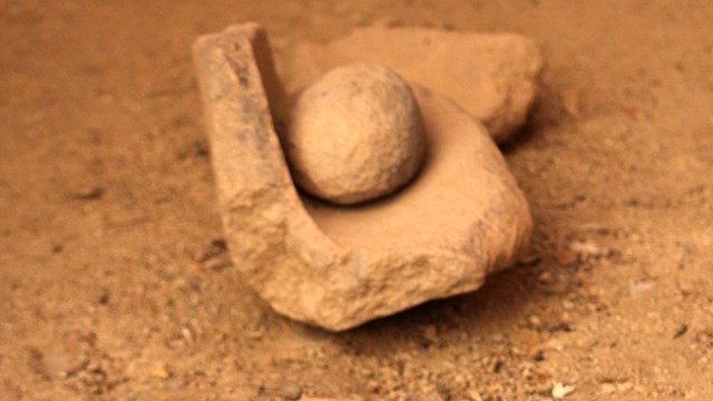 Mano and metate of orange stone. A round stone ball sits in the curve of a bowlish-shaped stone.