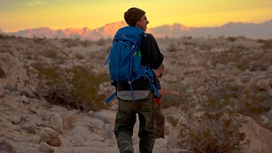 A hiker wearing a backpack on a trail at sunset.