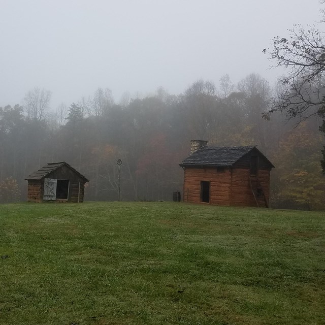 Two small log structures in an open pasture surrounded by trees. NPS photo. 