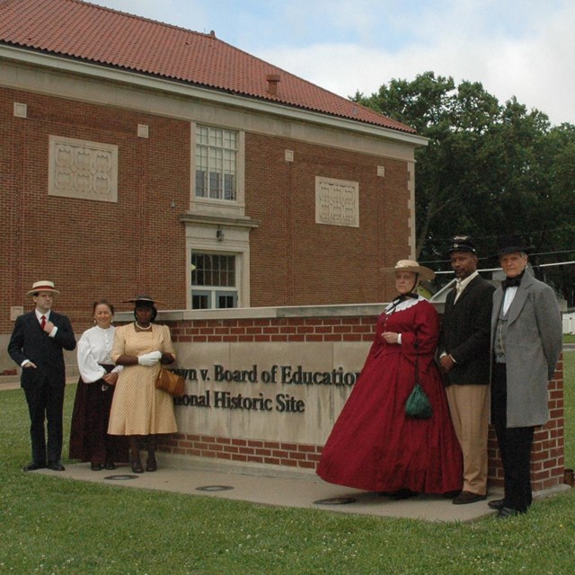 Characters in period dress gather around park entrance sign, brick building in rear. NPS photo. 