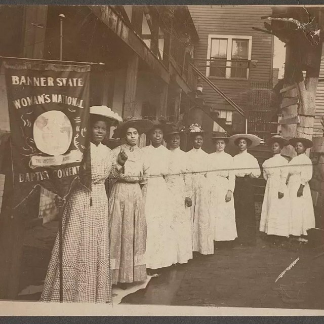 sepia photo of black women in victorian era dress holding a banner for a women's organization.