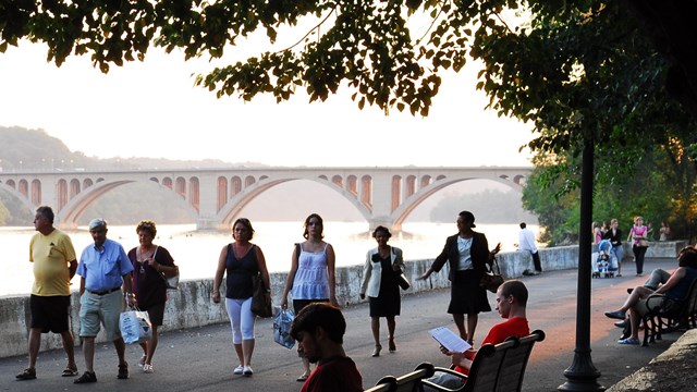 Group of people walking on path next to Potomac River. Francis Scott Key Bridge in background.