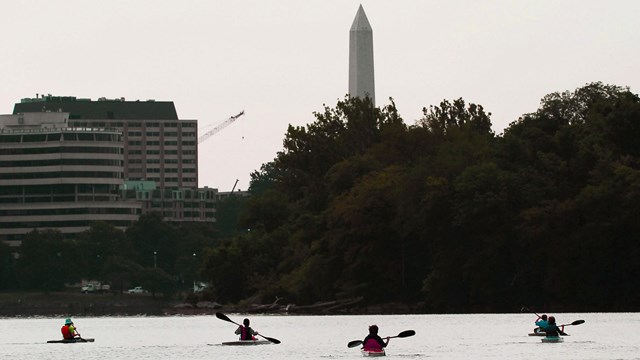 People in small boats row on Potomac River with Washington Monument behind him.