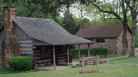 two log cabin structures, one with a brick chimney, green grass, framed with trees