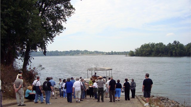 A group of people wait at the bottom of a landing along a river.