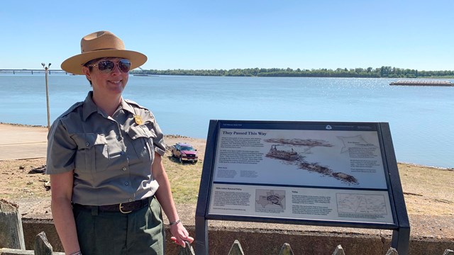 A uniformed park ranger stands next to a wayside exhibit along a river.