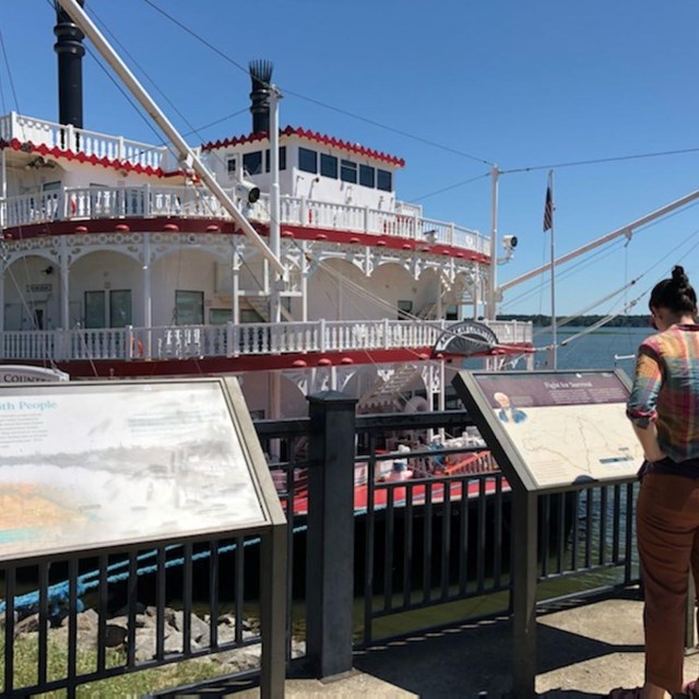 A person stands reading an exhibit sign in front of a white, historic steam ship.