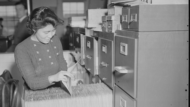 A Japanese American woman puts a file into a file shelf drawer full of file