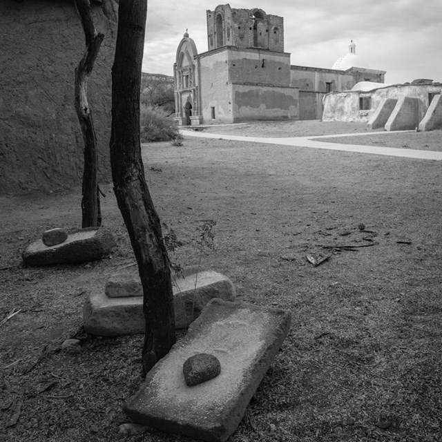 Black and white photograph of grinding stones on the ground with adobe church.