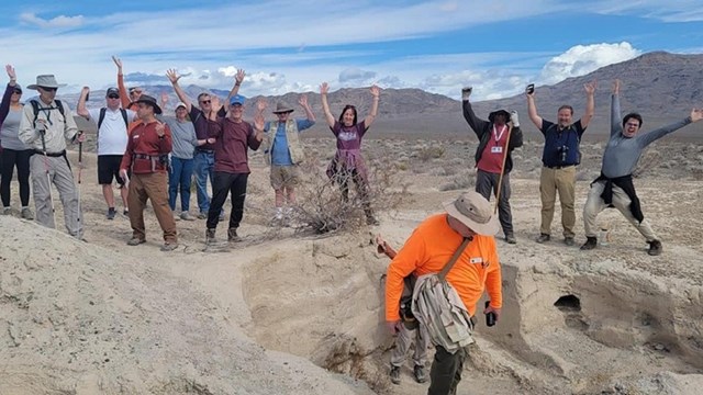 A group of visitors among tan badlands with their arms raised. 